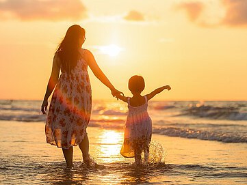 Mutter und Kind laufen Hand in Hand während dem Sonnenuntergang am Strand entlang.